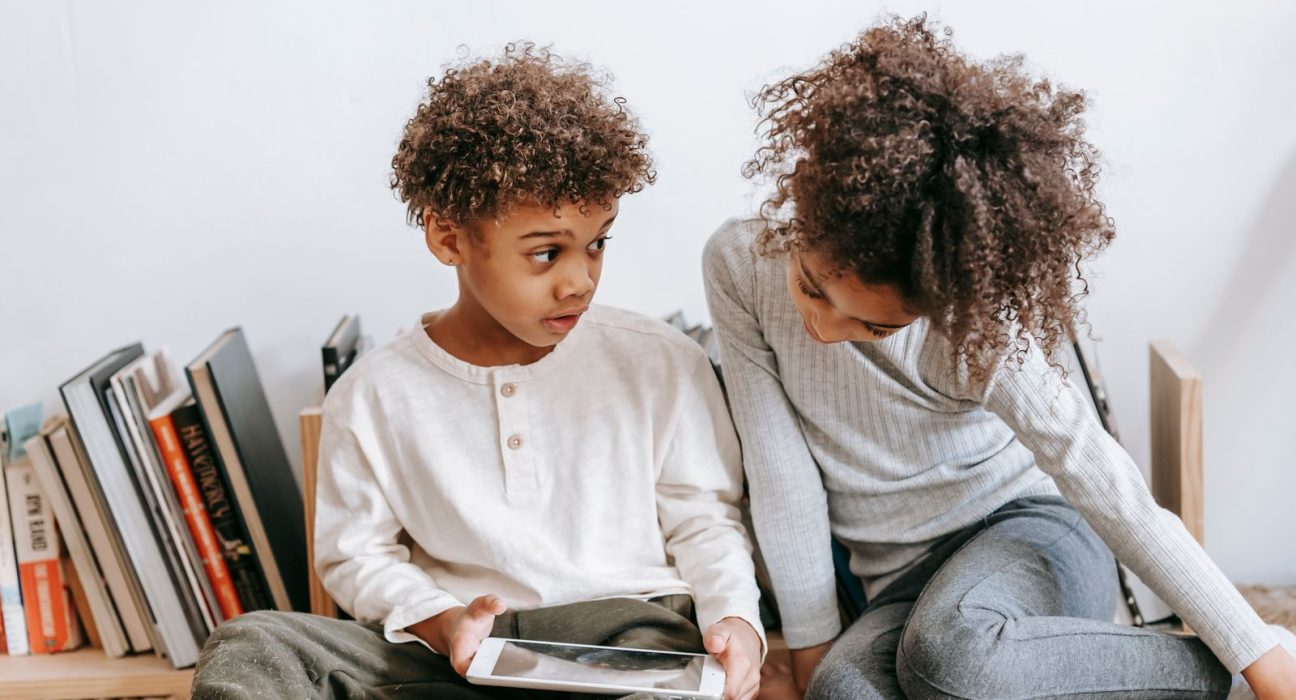 Two children sitting by a bookshelf, one holding a tablet