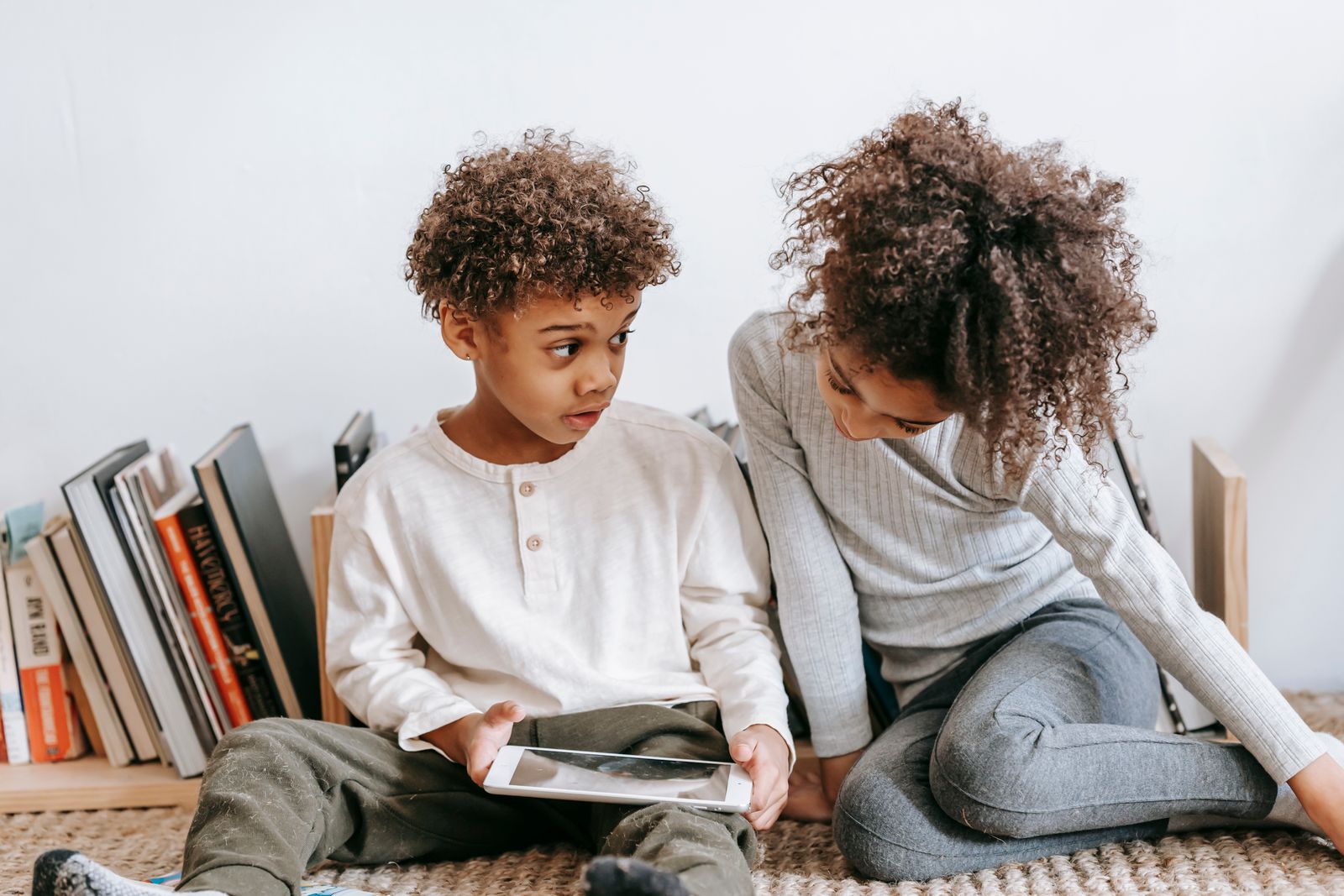 Two children sitting by a bookshelf, one holding a tablet